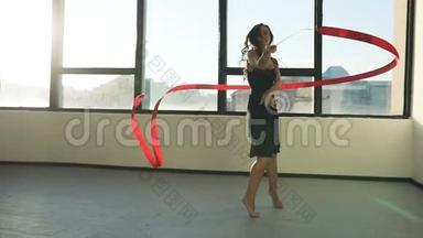布鲁内特年轻女子舞蹈<strong>艺术体操</strong>，现代用红色丝带穿黑色连衣裙在窗户前。 太阳